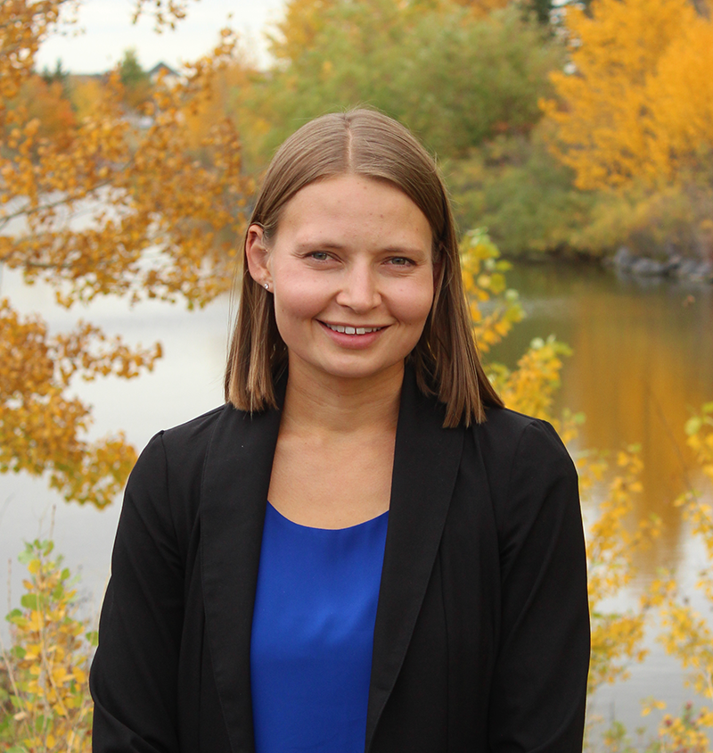Jill de Grood smiles at the camera wearing a black blazer with a blue shirt. She stands outside in front of a river with surrounding autumnal trees.
