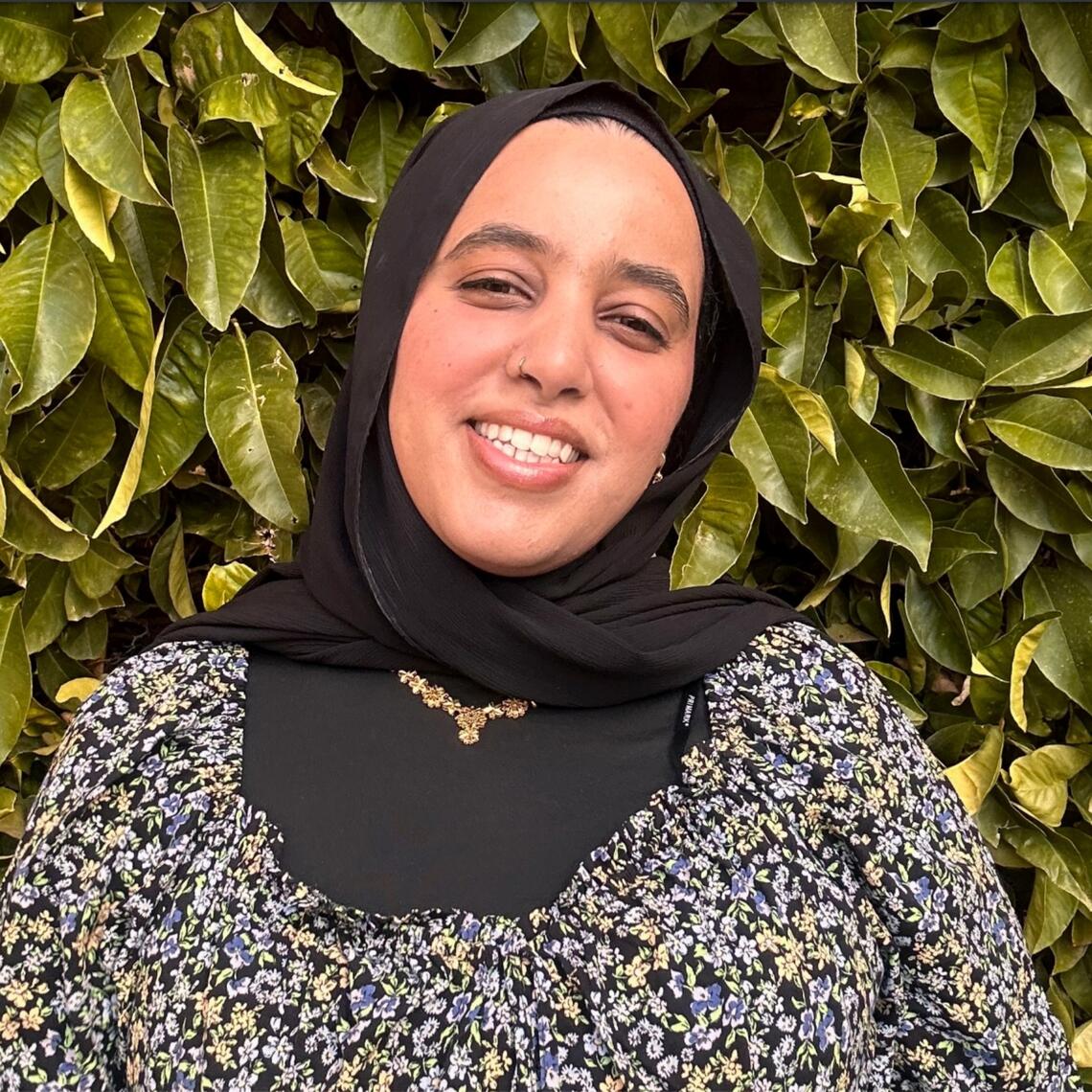 A woman in a black hijab and a white and black top smiles at the camera in front of green leaves