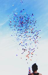 A person pops a confetti popper and confetti explodes into the air against a blue sky.
