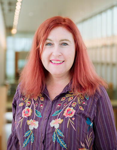 Kristen Gillespie-Lynch, a white woman with bright red hair, is wearing a purple embroidered shirt and smiling inside the Taylor Insitute for Teaching and Learning