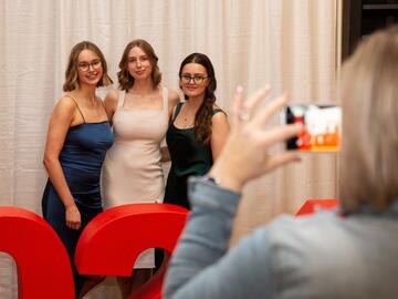Three women posing while an onlooker takes a photo on cell phone.